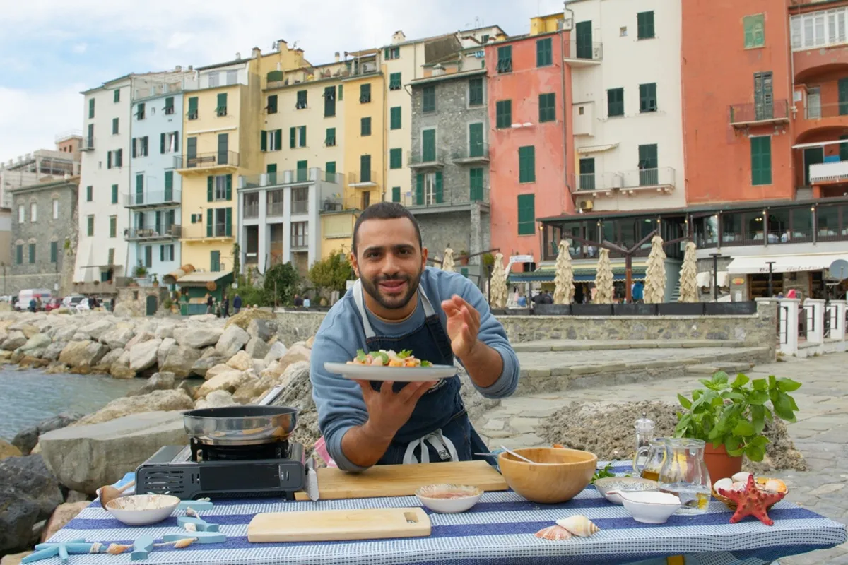 Ruben Bondì mostra con orgoglio un piatto di pesce fresco preparato all'aperto, con una tavola apparecchiata in riva al mare e case colorate sullo sfondo di un borgo costiero italiano.