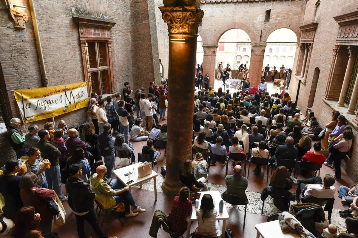 Auditório a céu aberto com dezenas de pessoas assistindo a um evento cultural em um pátio de arquitetura antiga com paredes de tijolos e colunas ornamentadas.