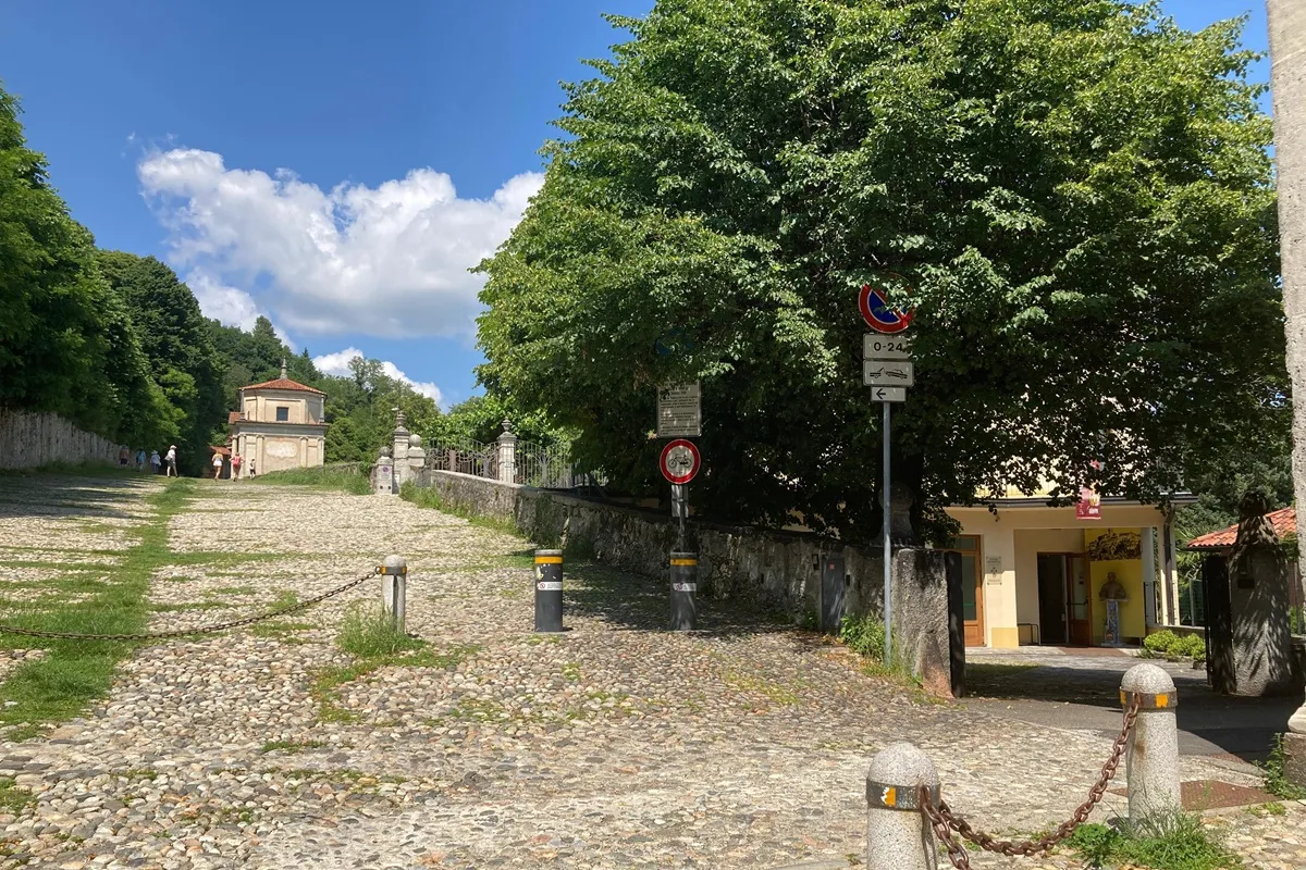 Caminho de pedras do Sacro Monte di Varese ladeado por árvores verdes, com capela ao fundo e céu azul com nuvens.