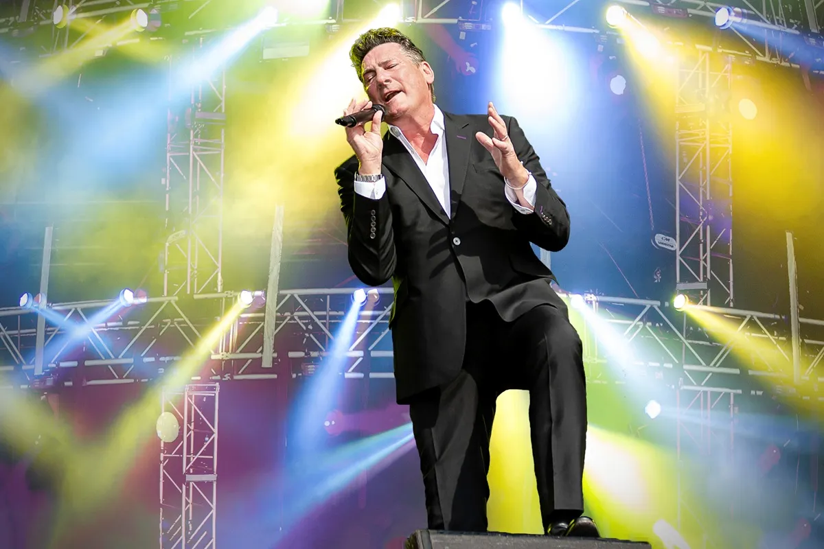 Tony Hadley canta ao vivo em um show da turnê “Tony Hadley 45”, vestido com terno preto, cercado por luzes coloridas e estrutura de palco iluminada.