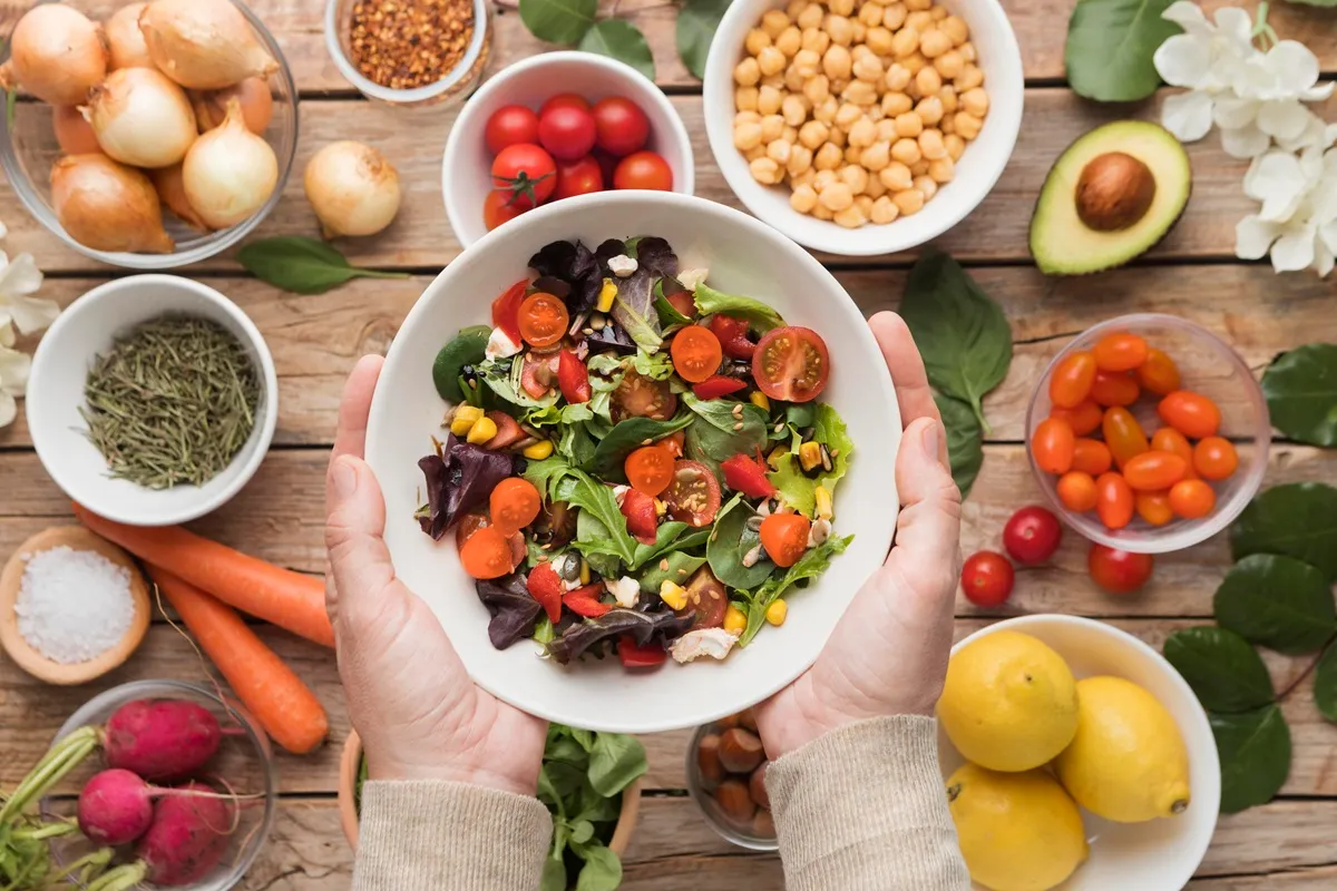 Ciotola di insalata mista con pomodorini, carote, mais e lattuga tenuta tra le mani, circondata da ingredienti freschi come ceci, avocado, limoni, cipolle e spezie