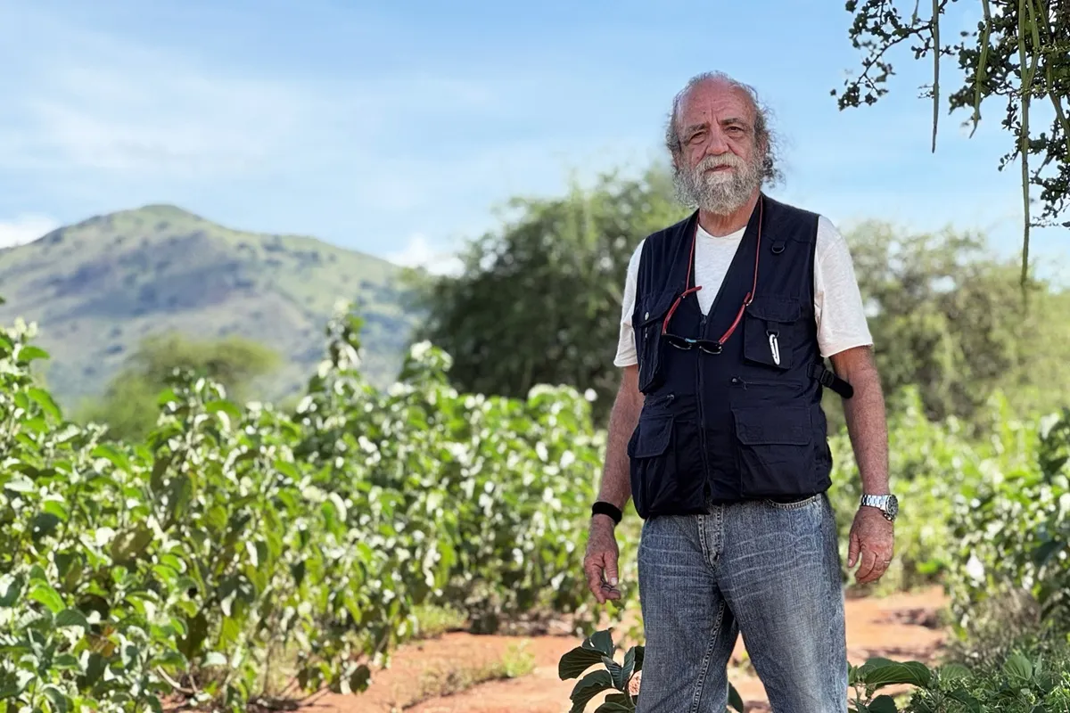 Uomo anziano con barba bianca e capelli grigi, che indossa un gilet da fotografo, in un campo con piante verdi e montagne sullo sfondo, in Kenya