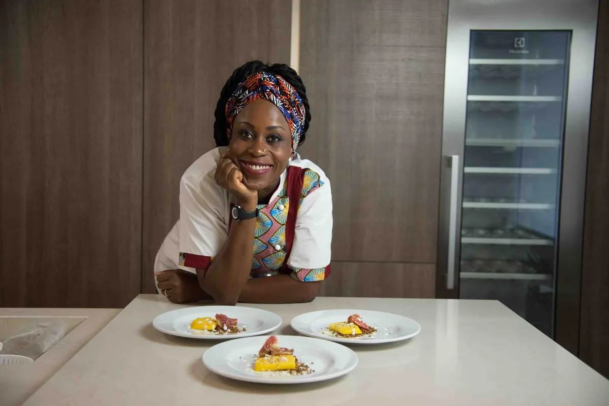 Chef Victoire Gouloubi sorride appoggiata al bancone con tre piatti gourmet afro-caraibici davanti a sé, in una cucina moderna.