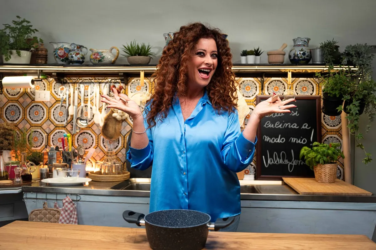 Valentina Persia sorrindo e fazendo um gesto divertido na cozinha, com fundo colorido e detalhes de plantas e utensílios culinários.