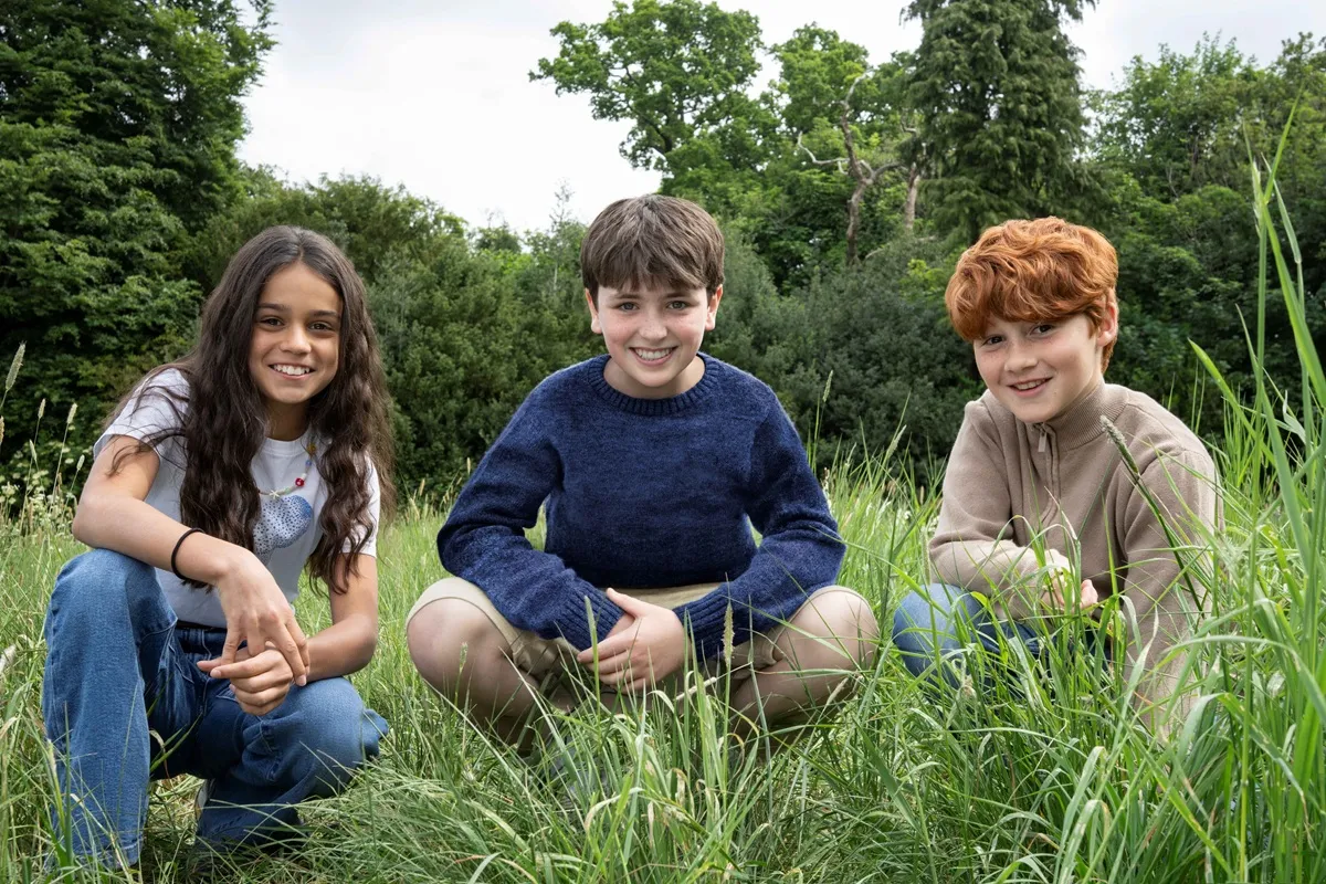 Dominic McLaughlin, Arabella Stanton e Alastair Stout seduti in un prato, sorridendo, scelti come Harry Potter, Hermione Granger e Ron Weasley nella nuova serie HBO