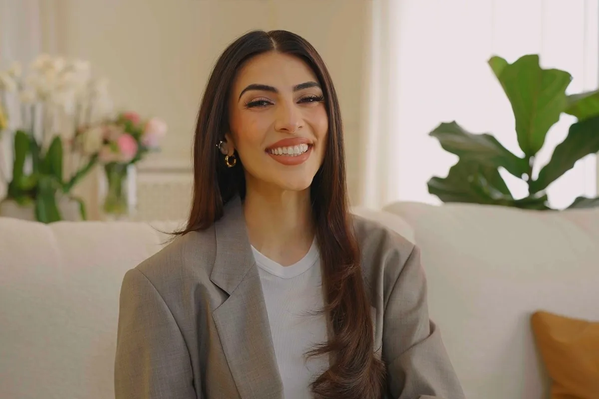 Donna sorridente con capelli lunghi e castani, indossa blazer grigio e orecchini dorati, seduta su divano in ambiente elegante
