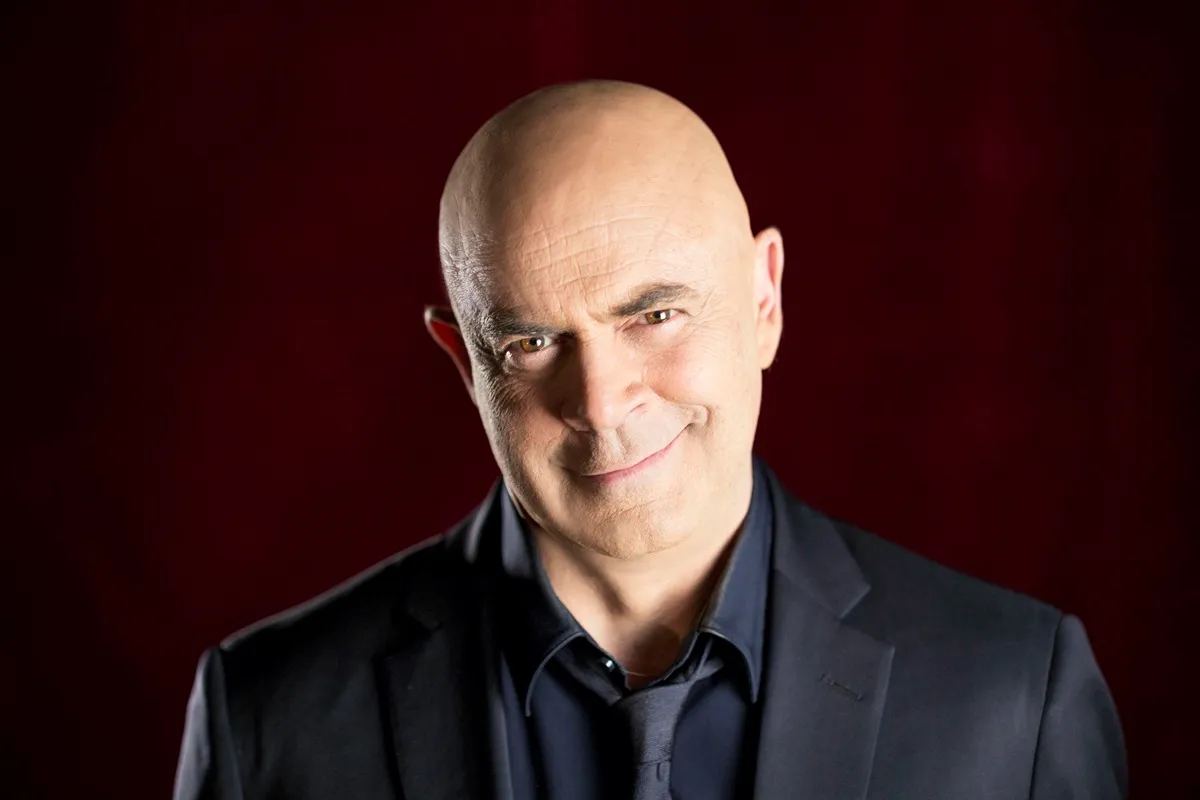 Immagine di Maurizio Crozza, il comico genovese, protagonista del programma "Fratelli di Crozza". L'artista sorride con uno sguardo malizioso, indossando un completo elegante, con un sfondo rosso scuro.