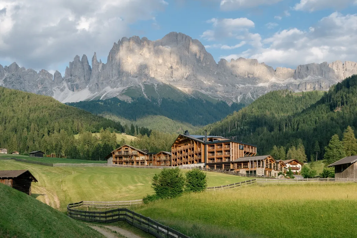 Veduta di un resort in legno immerso nei prati verdi ai piedi delle Dolomiti, con le maestose vette del gruppo del Catinaccio sullo sfondo