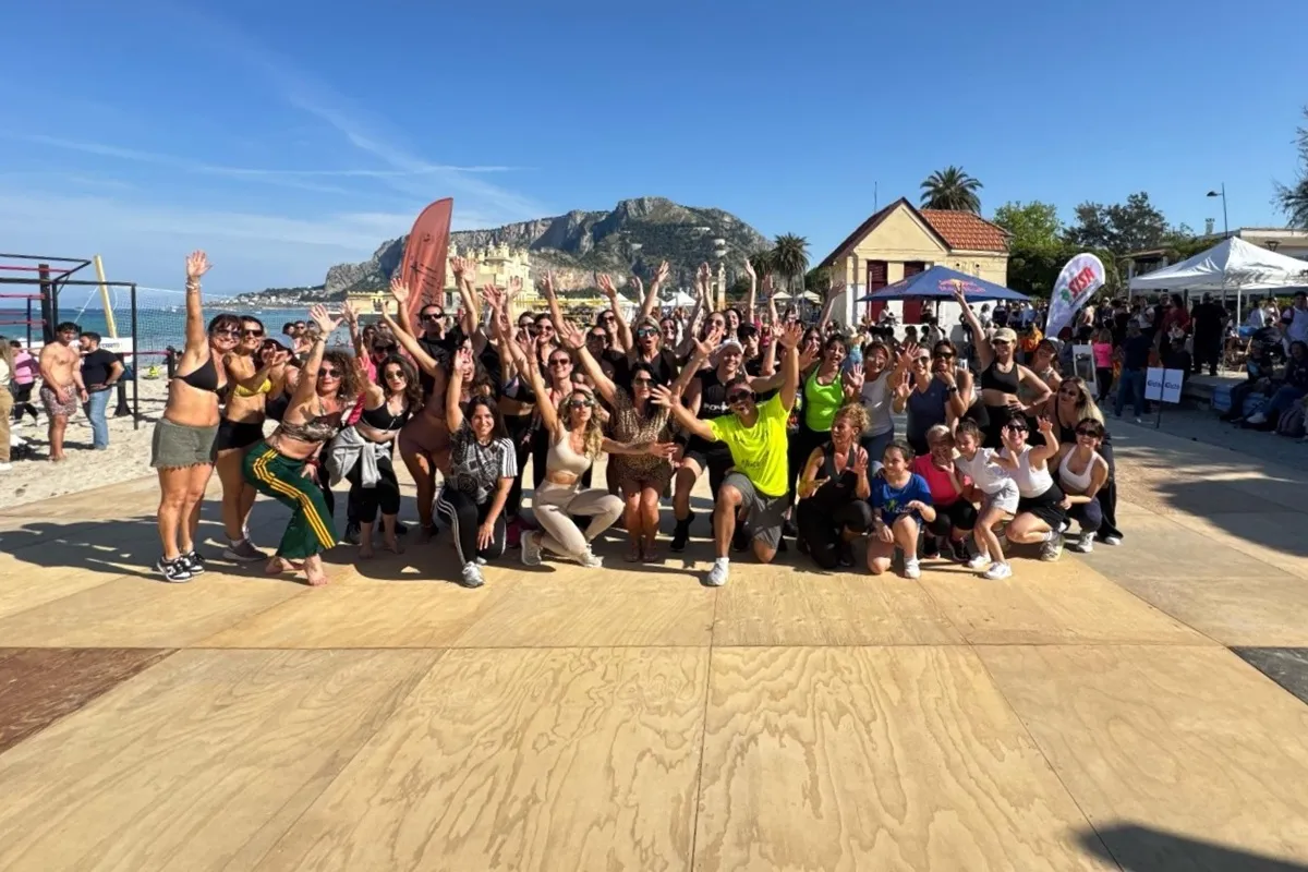 Gruppo di persone sorridenti che partecipano a un evento fitness sulla spiaggia di Mondello, con il mare e le montagne sullo sfondo
