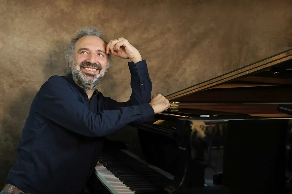 Stefano Bollani sorride seduto al pianoforte a coda, indossando una camicia scura, con sfondo marrone neutro