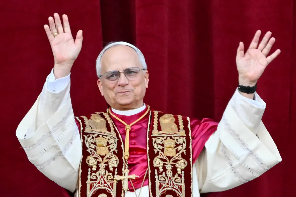 Cardinale del Vaticano in abito liturgico rosso e dorato, saluta sorridendo con una mano alzata durante un evento religioso.