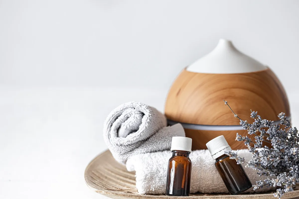 Diffusore di aromaterapia in legno, bottiglie di oli essenziali e asciugamani bianchi su un vassoio, con un ramo di lavanda decorativo.