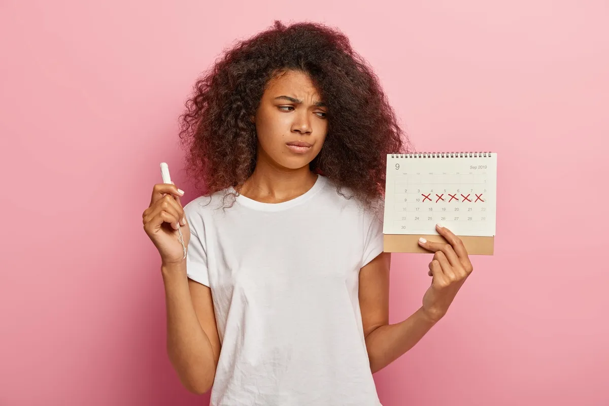 Giovane donna perplessa con tampone in mano che osserva un calendario segnato con croci rosse per monitorare il ciclo mestruale
