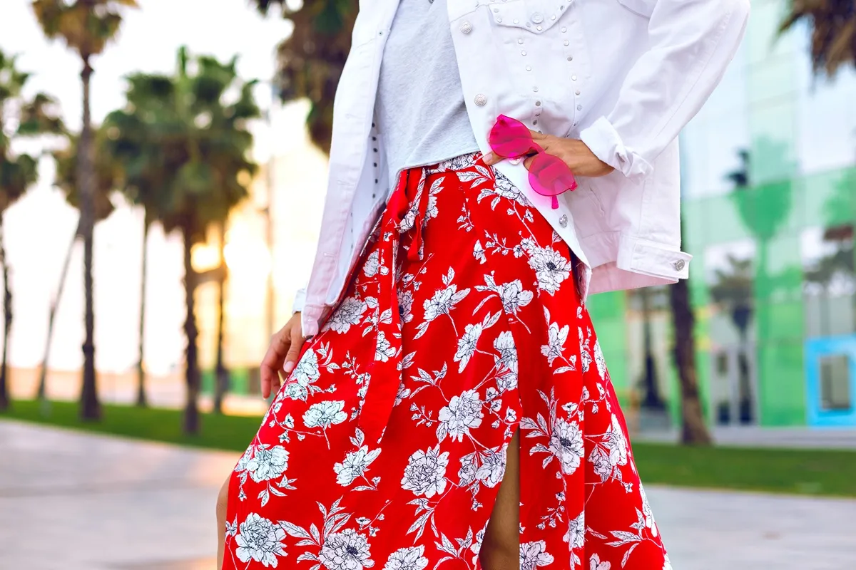 Dettaglio di outfit estivo con gonna lunga rossa a fiori bianchi, giacca bianca e occhiali da sole rosa