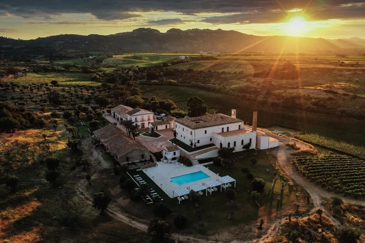 Immagine aerea di una vigna con vista sulle montagne sullo sfondo, evidenziando una grande villa in stile tradizionale, con un ampio giardino e piscina al centro. Il tramonto illumina delicatamente la scena, creando un ambiente tranquillo e accogliente.