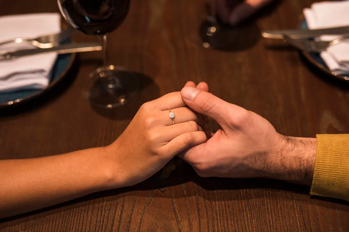 Coppia che si tiene per mano al ristorante, con l'anello di fidanzamento visibile sulla mano della donna, in un ambiente intimo e romantico.