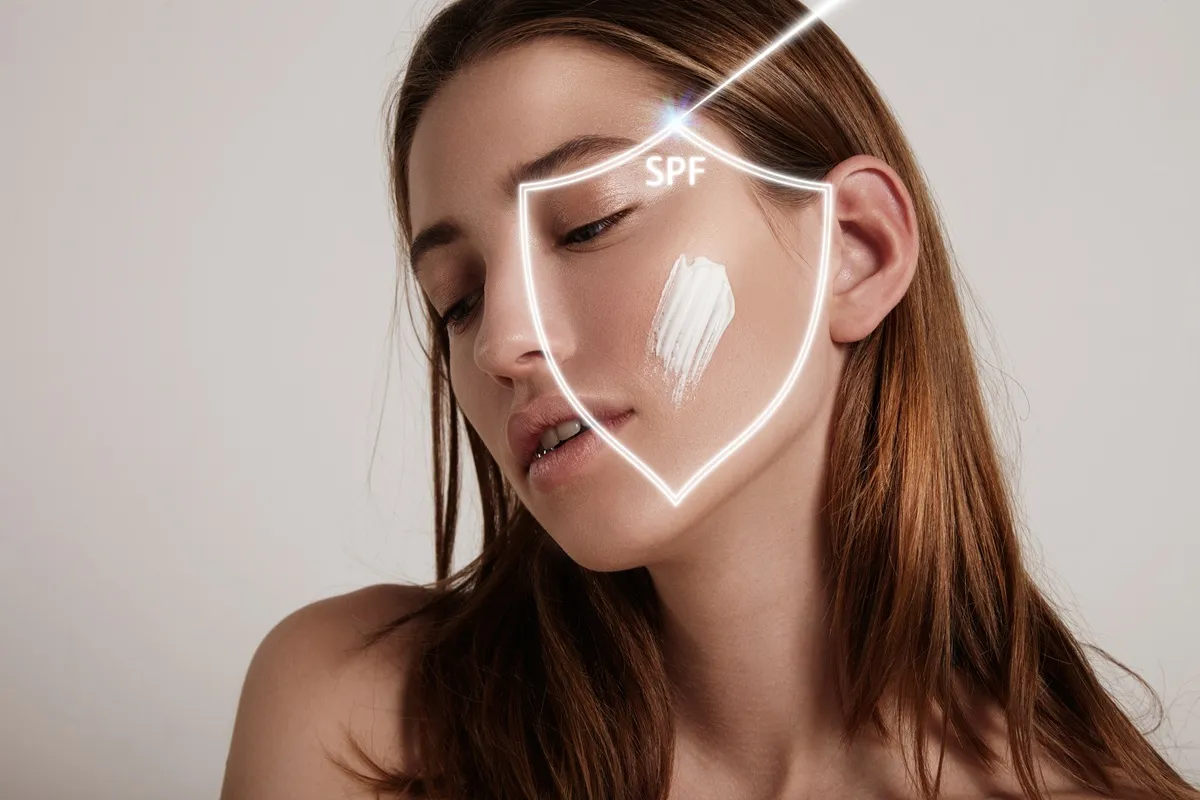 Immagine di una donna che applica una crema solare sul viso con un'indicazione visibile di SPF per protezione dai raggi UV.
