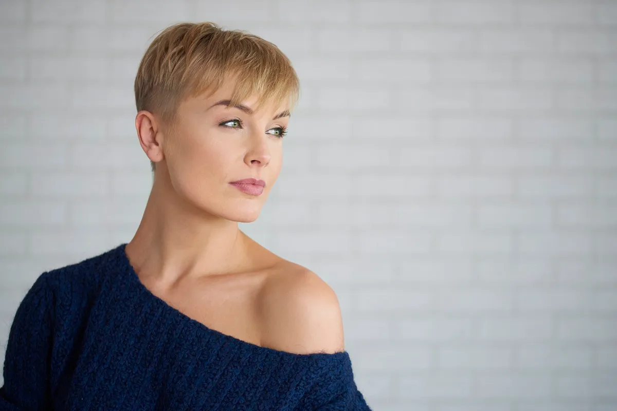 Donna con taglio di capelli cortissimo, indossa un maglione blu scuro e guarda in lontananza con espressione riflessiva.