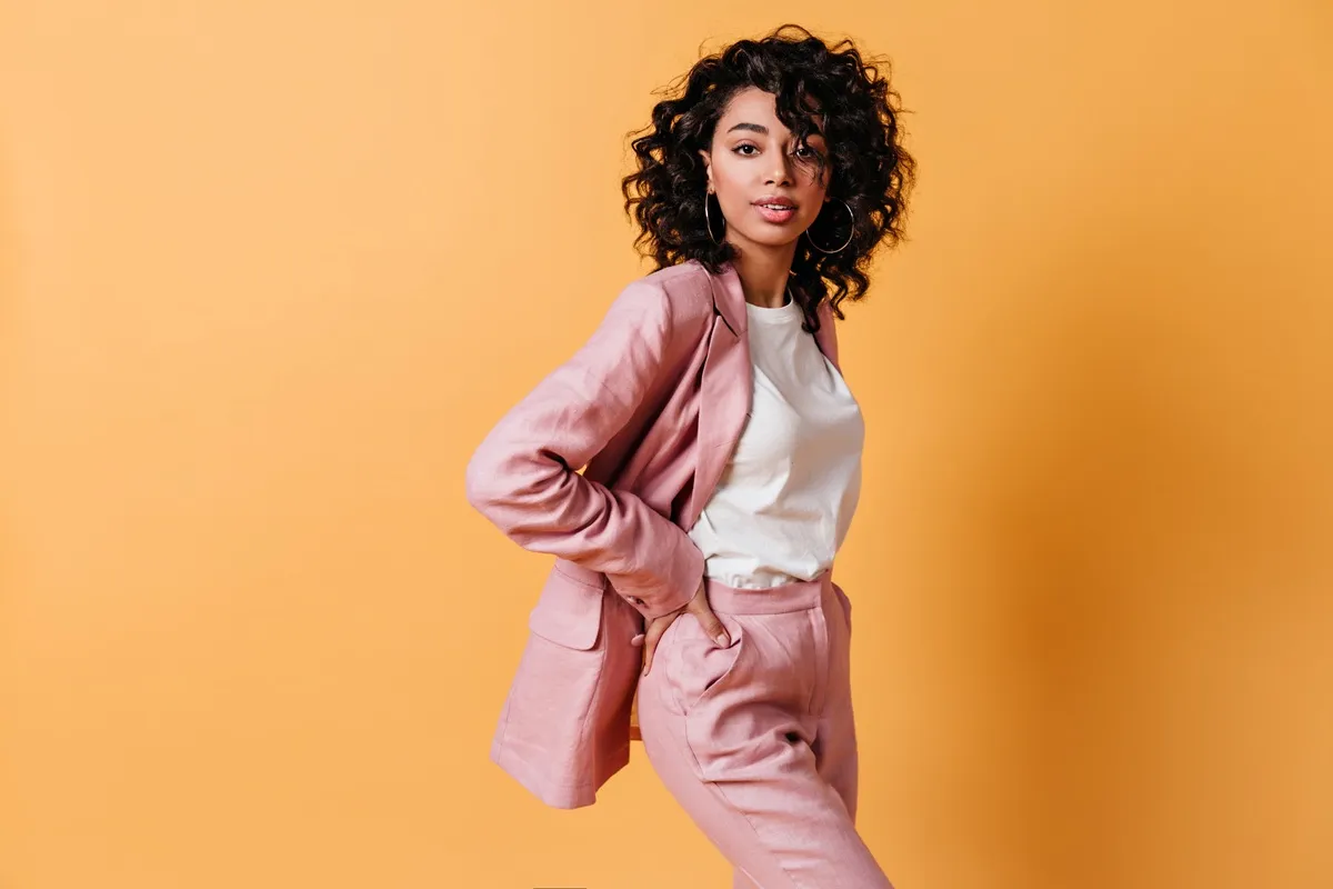 Donna con capelli ricci indossa un tailleur rosa con t-shirt bianca davanti a sfondo arancione