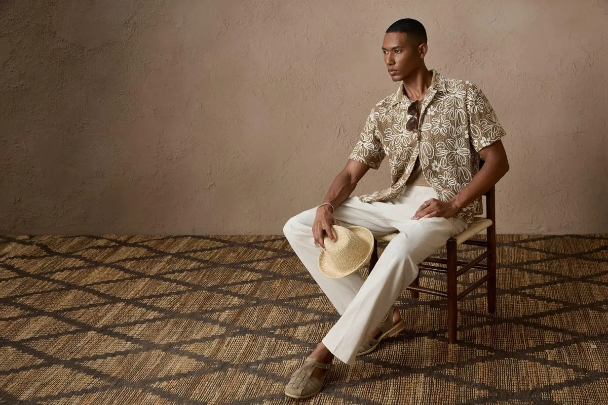 Modello in posa con camicia hawaiana e pantaloni chiari, seduto su una sedia con un cappello in mano