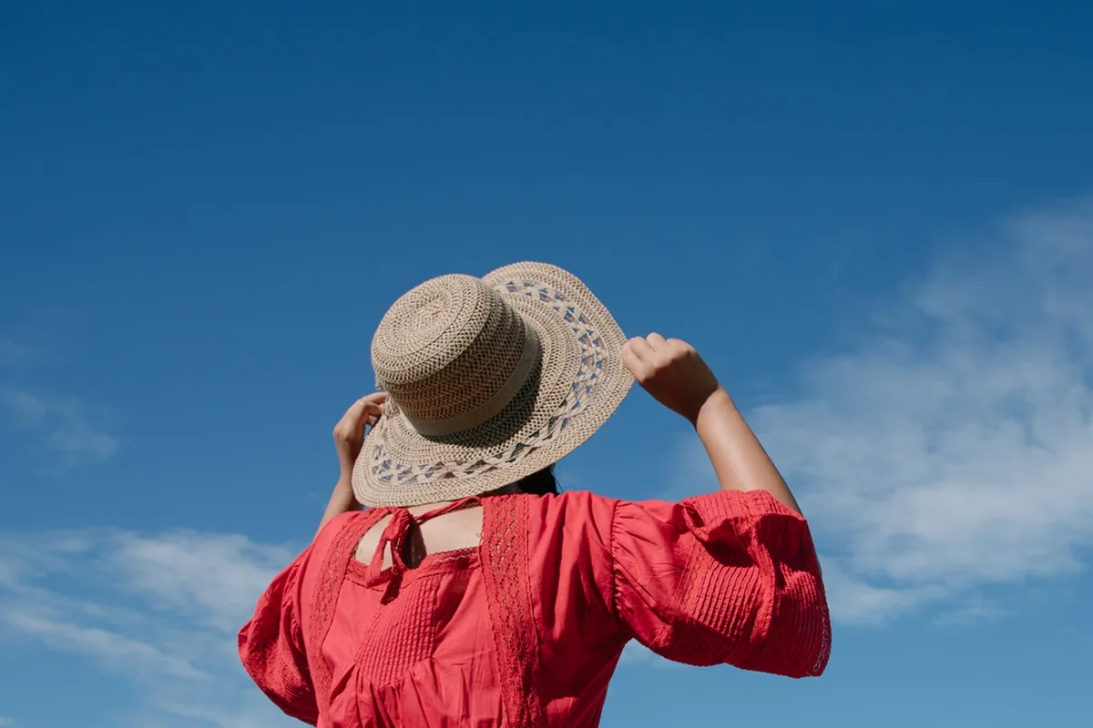 Donna che indossa un vestito rosso e un cappello di paglia, guardando il cielo azzurro e sorridendo.