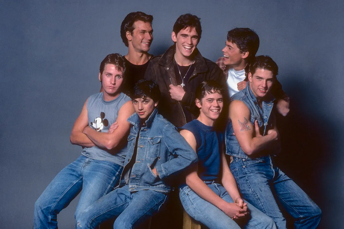 Gruppo di sei giovani uomini in abiti anni ’80 (jeans e giacche di jeans), sorridenti e in posa su sfondo neutro, foto promozionale di The Outsiders