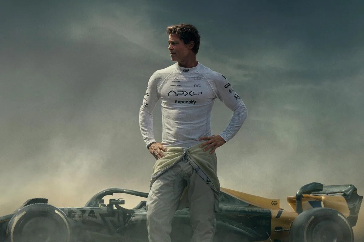 Brad Pitt nei panni del pilota Sonny Hayes in tuta bianca, in piedi accanto a un’auto di Formula 1 con il logo Apx GP sul cofano, sul set di F1 – Il Film