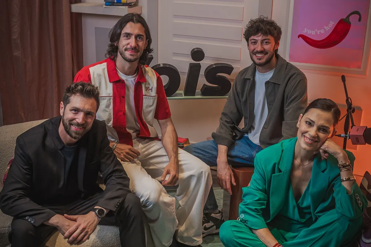 Foto dei presentatori dei video podcast di Vois TV, sorridenti e posati insieme nel loro studio. Si notano elementi visivi moderni e colorati nello sfondo, con il logo Vois ben visibile.