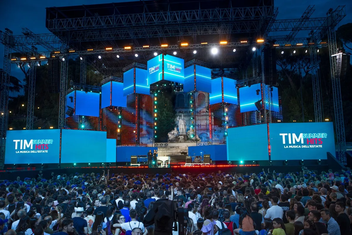 Immagine del palco del TIM Summer Hits 2025, con luci colorate e una grande folla che assiste al concerto in Piazza del Popolo a Roma. Il palco è decorato con schermi LED e il logo dell'evento.