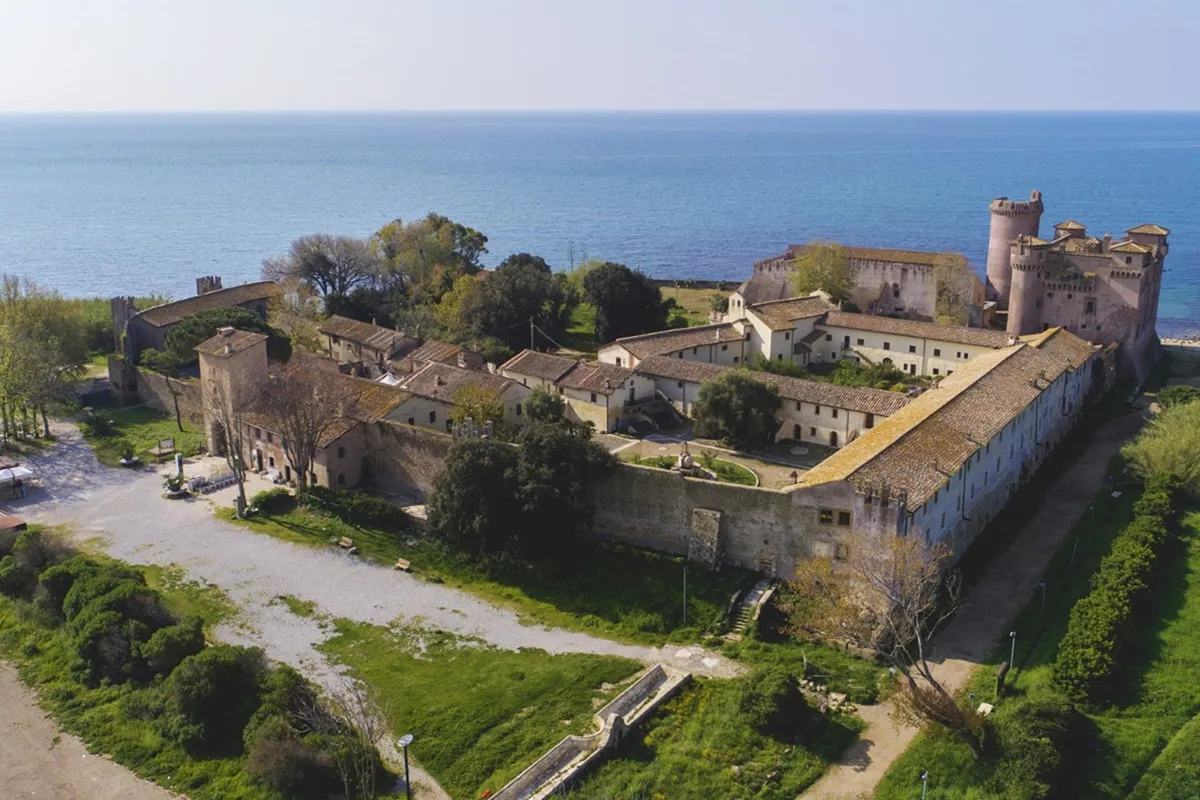 Veduta aerea del Castello di Santa Severa affacciato sul Mar Tirreno, con mura medievali, cortile interno e ampio prato.