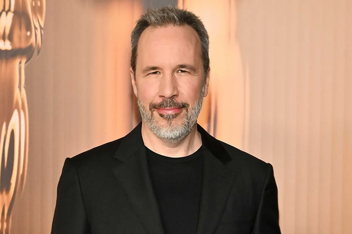 Denis Villeneuve in giacca nera, mezzo busto, sorride alla 97ª Nominees Dinner degli Academy Awards 2025 con statuetta dell’Oscar sfocata sullo sfondo dorato.