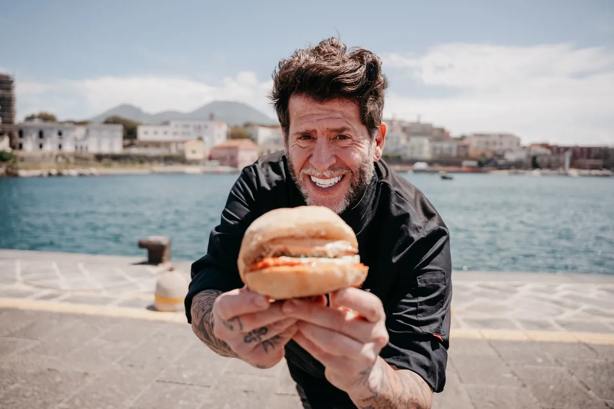 Vittorio Gucci, presentatore del programma "Il Panino Perfetto - Edizione Estiva", sorride mentre tiene un panino fresco, con il mare e la città sullo sfondo.