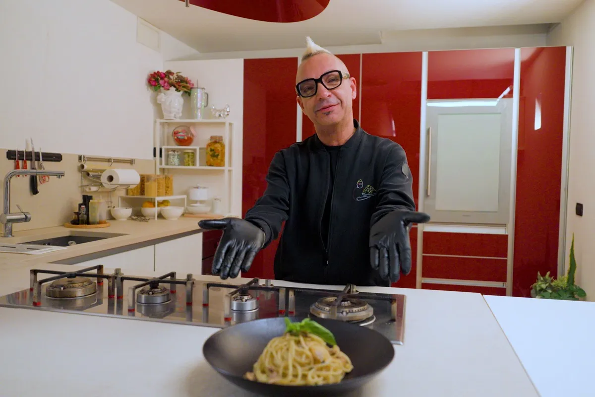 Angelo Roccavilla indossa guanti da cucina e presenta un piatto di pasta gourmet in un ambiente moderno e luminoso, tipico della sua cucina televisiva.