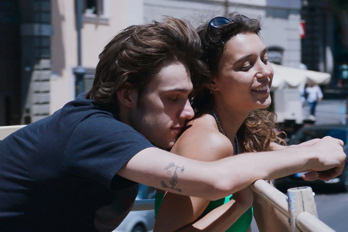 Una scena di Suspicious Minds con due giovani protagonisti, abbracciati e sorridenti mentre godono di un momento di intimità in una città affascinante.