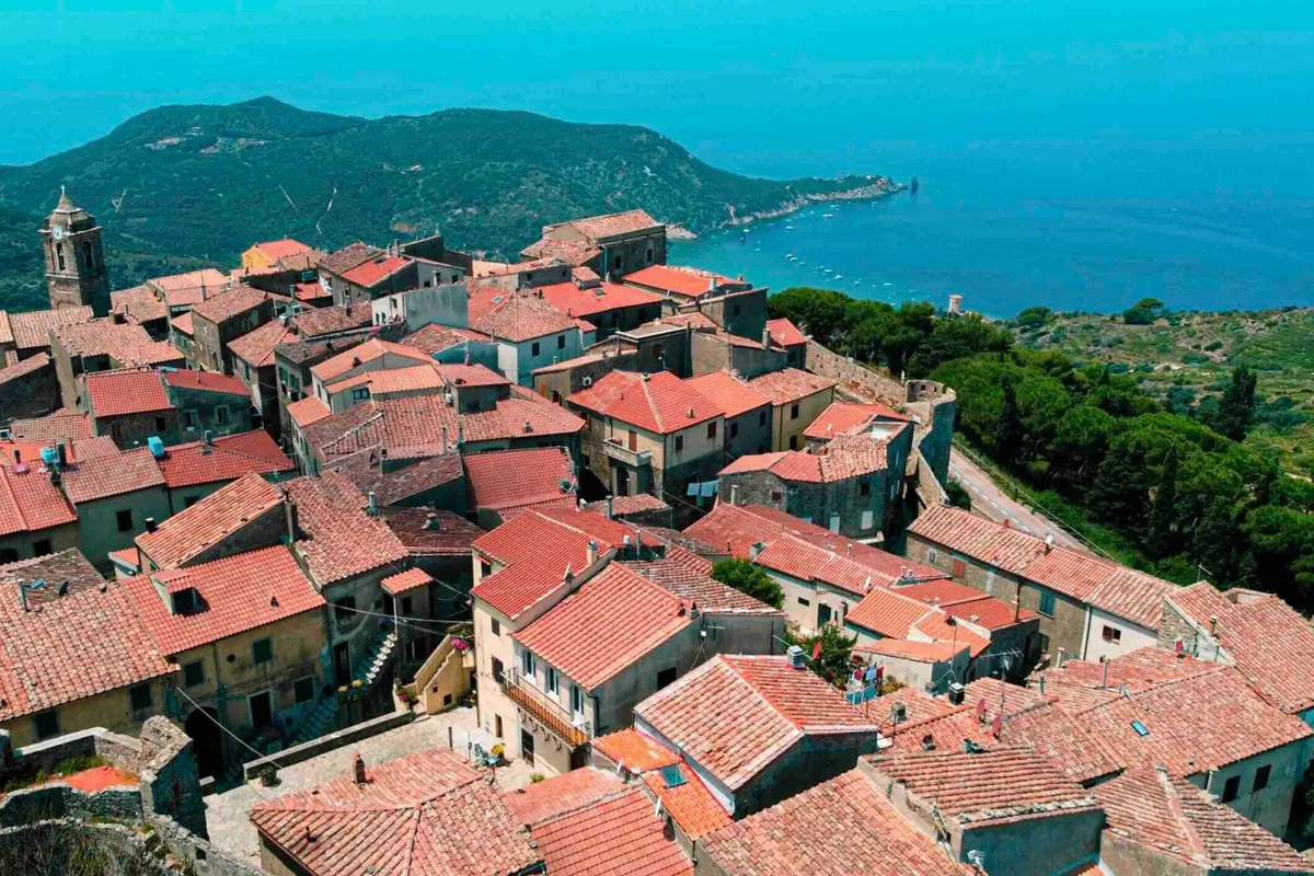 Veduta aerea del borgo di Giglio Castello su un promontorio roccioso, tetti in cotto e mare turchese sullo sfondo.