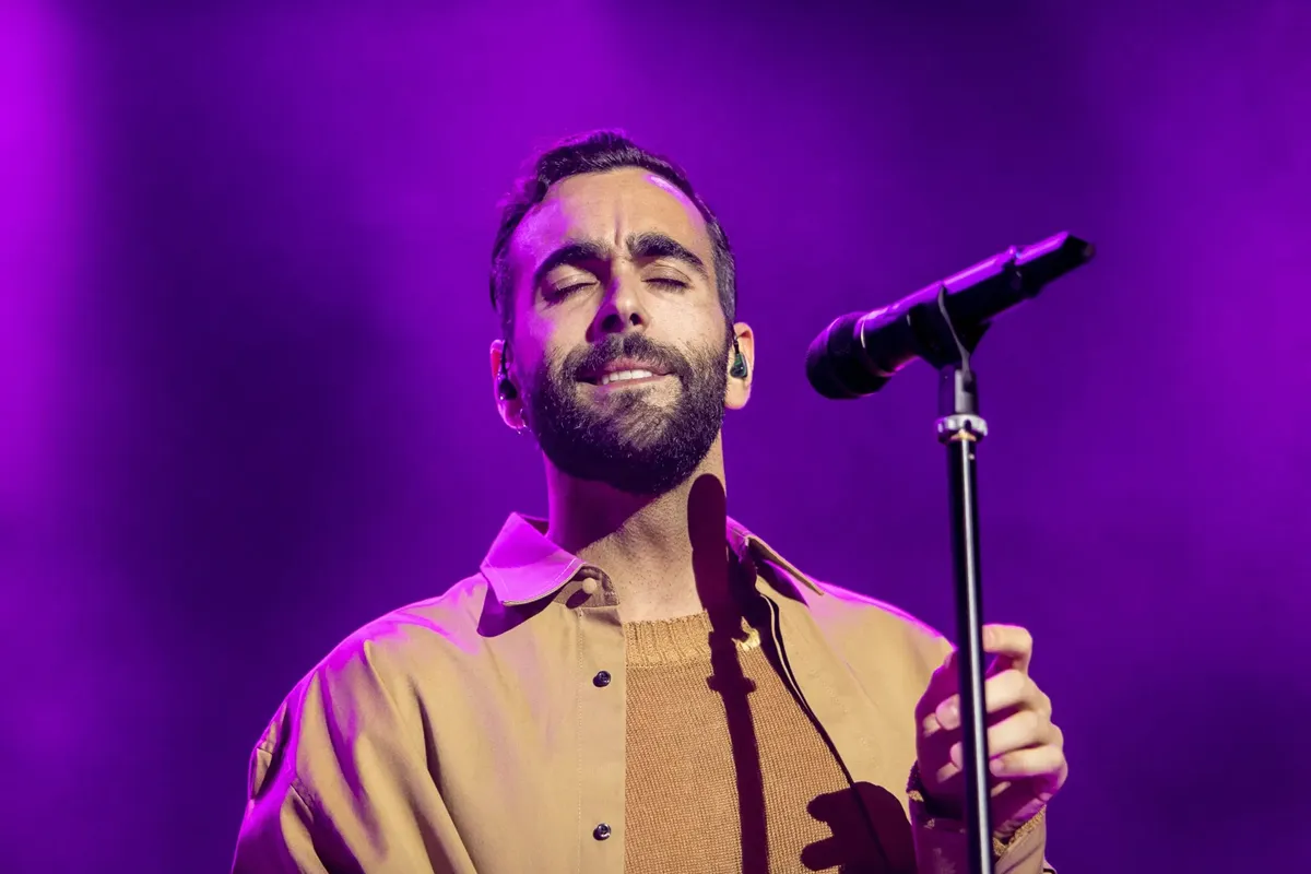Marco Mengoni canta durante un concerto a Zurigo, con illuminazione viola e un'espressione emozionata, indossando una camicia beige e tenendo un microfono.