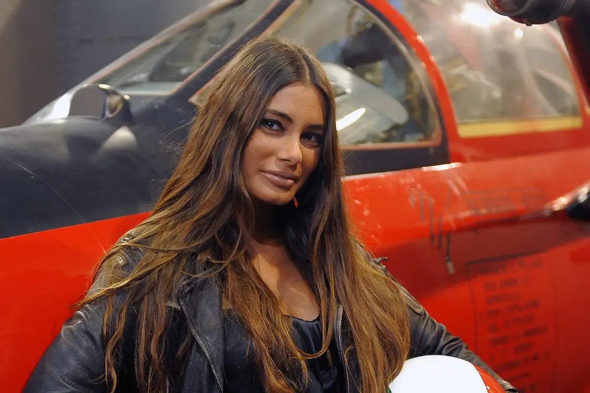 Donna bruna con lunghi capelli sciolti indossa una giacca di pelle nera mentre posa sorridente davanti a un aereo rosso.