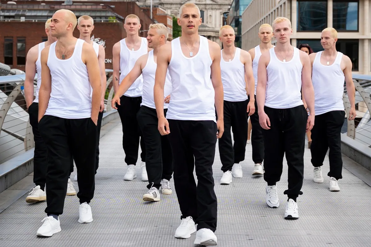 Un gruppo di uomini con capelli biondi e rasati cammina lungo un ponte a Londra, indossando magliette senza maniche bianche, pantaloni neri e scarpe bianche, con espressioni determinate.