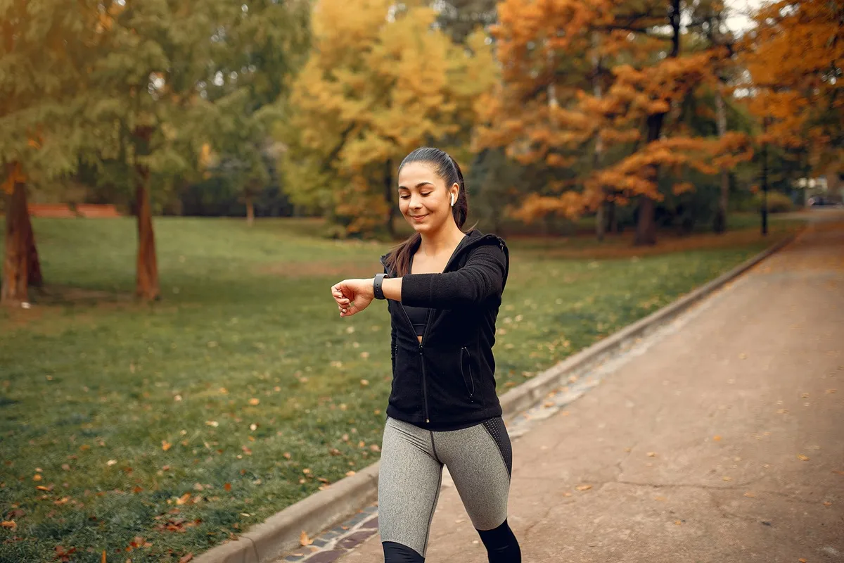 Immagine di una donna che cammina al parco mentre guarda l'orologio fitness al polso, con auricolari, indossando abbigliamento sportivo