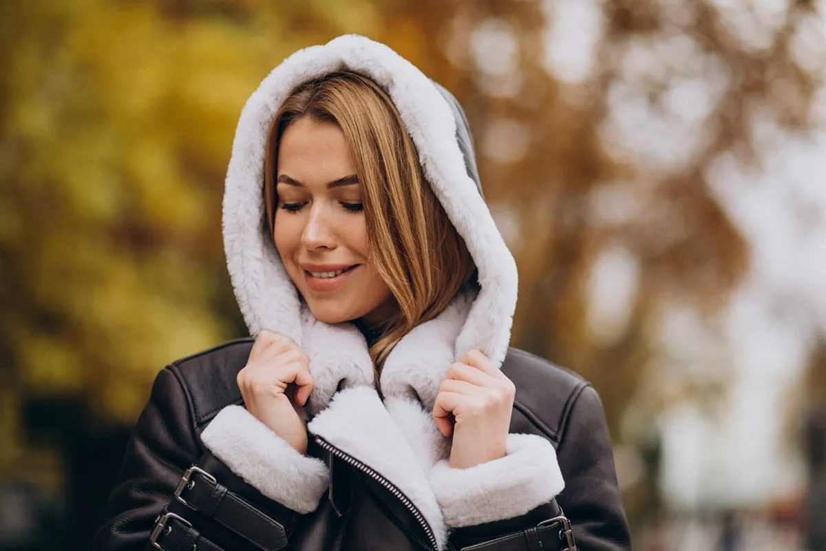 Donna con cappotto in pelle e interno in shearling bianco, cappuccio in testa, sorridente in esterno autunnale