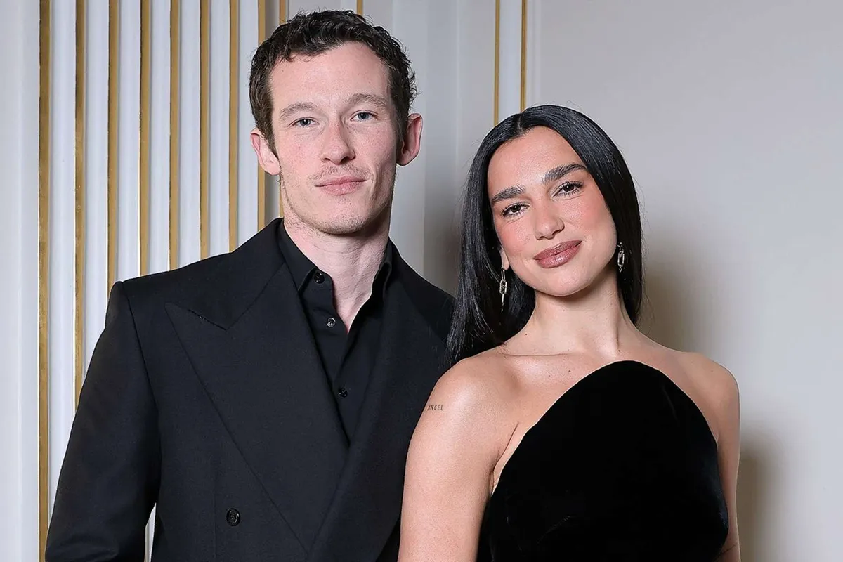 Dua Lipa e Callum Turner sorridono durante un evento pubblico, indossando abiti eleganti, con Dua in un abito nero senza spalline e Callum in un completo scuro.