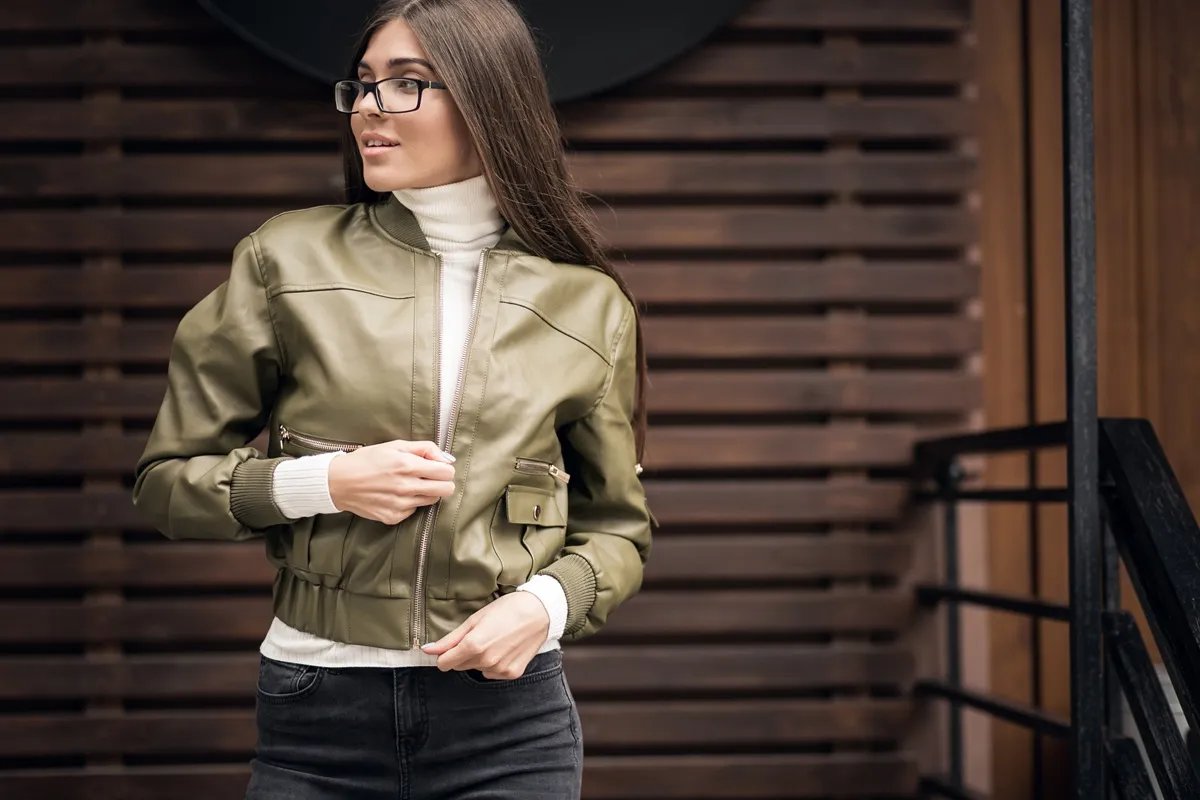 Donna con occhiali indossa giacca di pelle verde militare slim fit, abbinata a dolcevita bianco e jeans scuri, in ambiente urbano moderno
