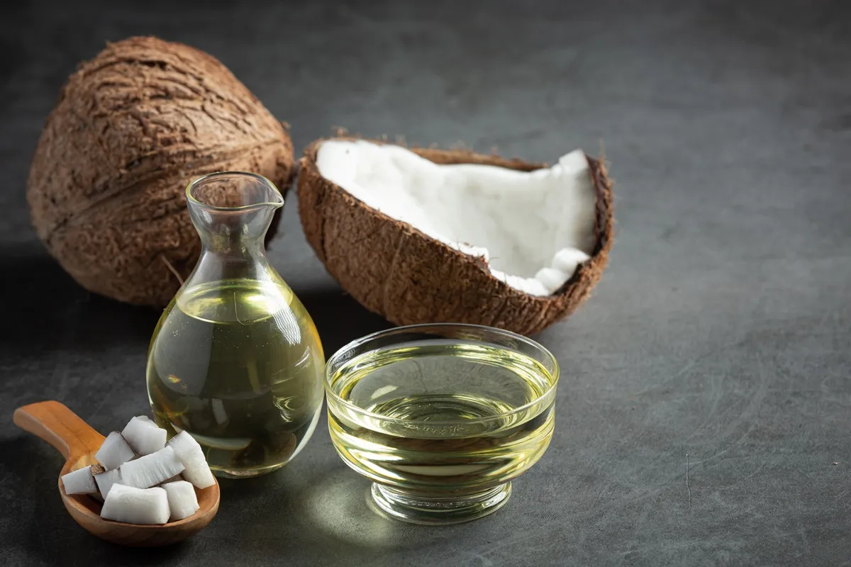 Immagine di olio di cocco in una ciotola e una bottiglia, con cubetti di polpa fresca di cocco e noci di cocco aperte sullo sfondo.