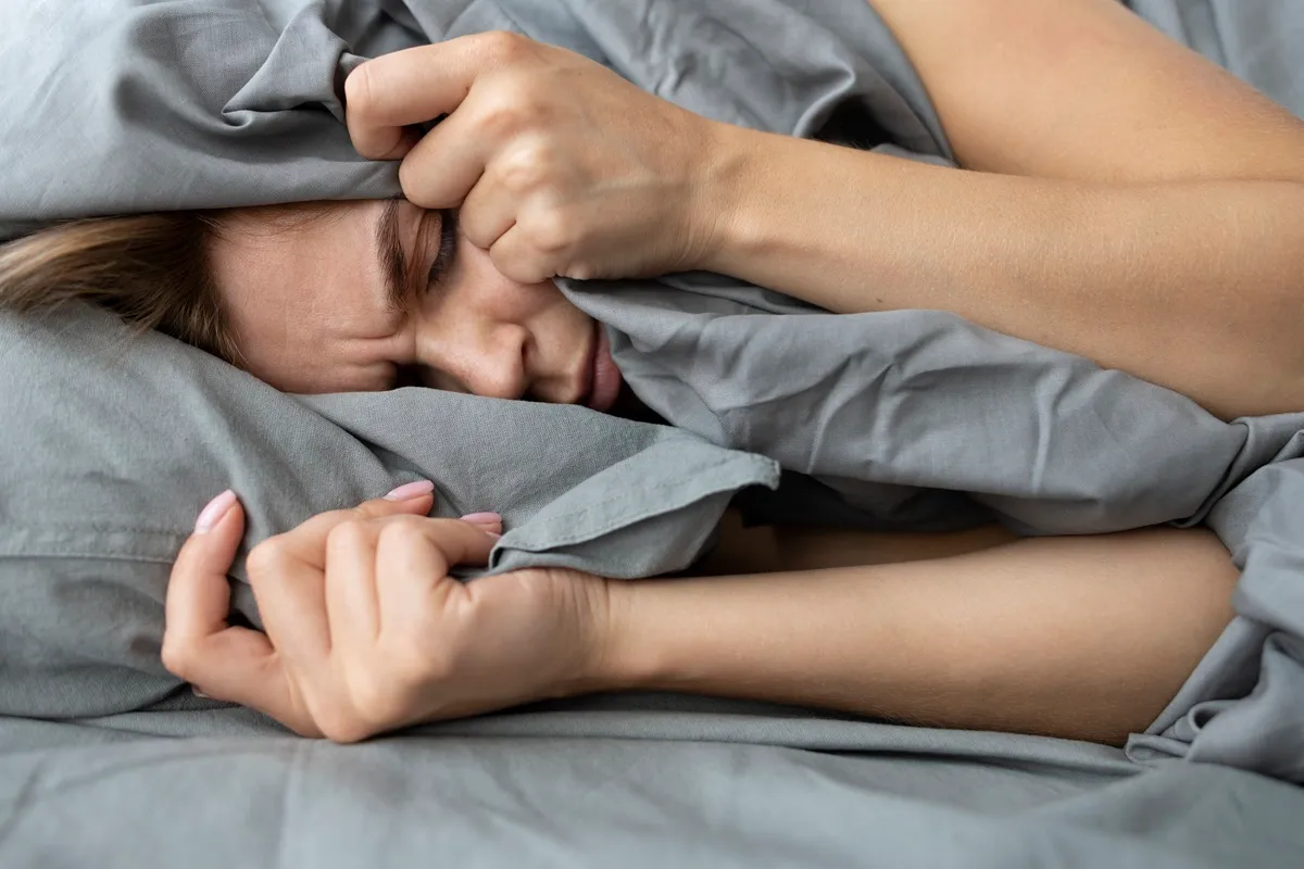 Immagine di una donna che soffre di paralisi del sonno, con il viso nascosto tra le coperte grigie, le mani che si stringono sul lenzuolo mentre manifesta disagio.