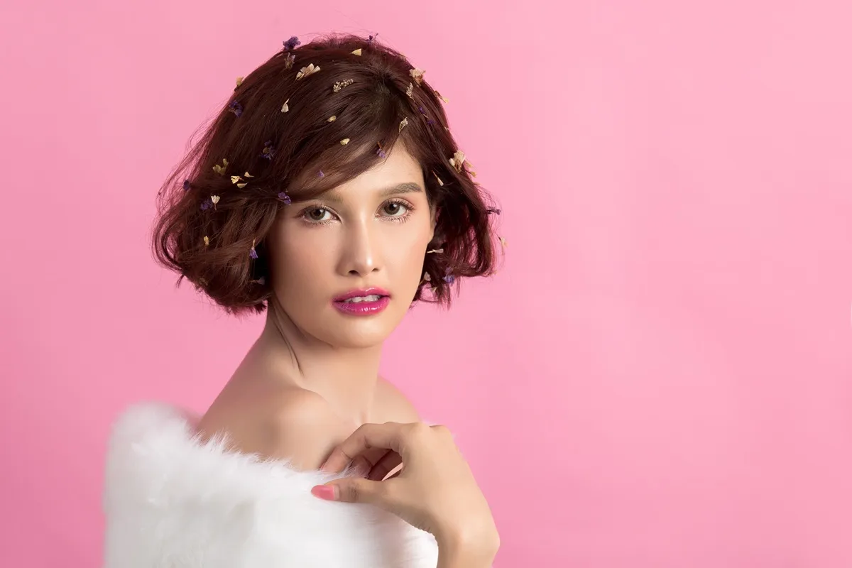 Ritratto di una giovane donna con capelli corti decorati con fiori, indossando una pelliccia bianca, con uno sfondo rosa.