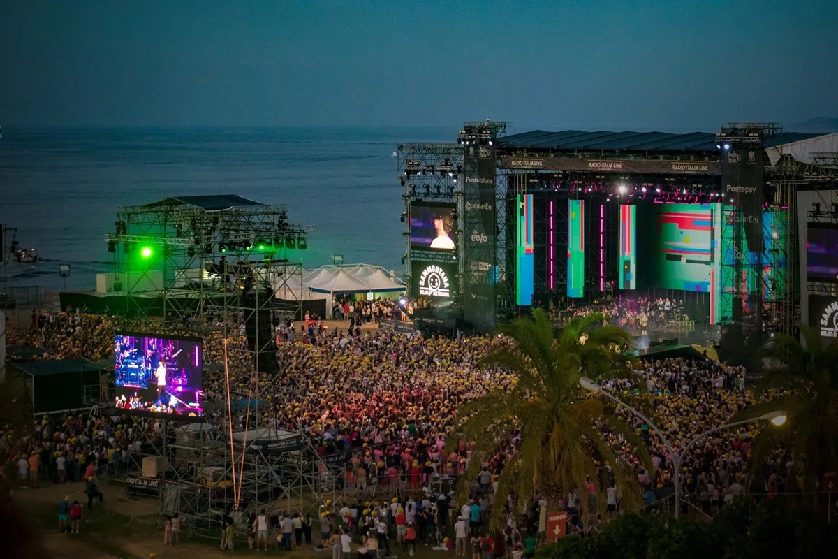 Palco illuminato del Radio Italia Live al Foro Italico di Palermo al tramonto, con grandi schermi LED, strutture metalliche, una folta platea di spettatori e vista sul mare e palme in primo piano