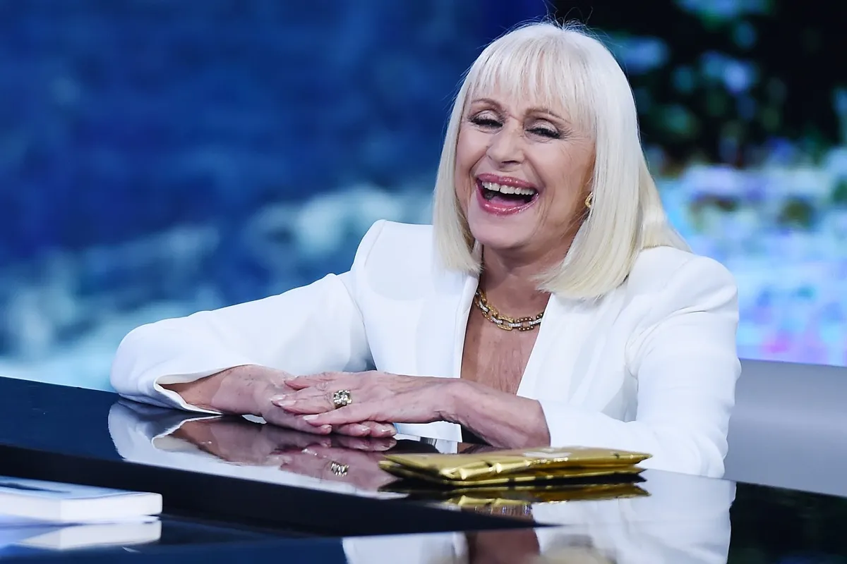 Raffaella Carrà con sorriso aperto, capelli biondo platino e blazer bianco, appoggiata al bancone di uno show televisivo.