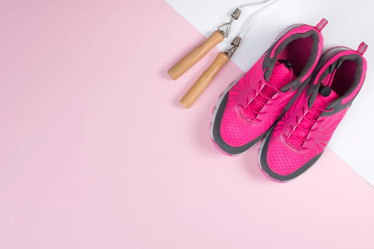 Scarpe da ginnastica rosa e grigie da donna affiancate a una corda per saltare con manici in legno, su sfondo rosa chiaro e bianco.