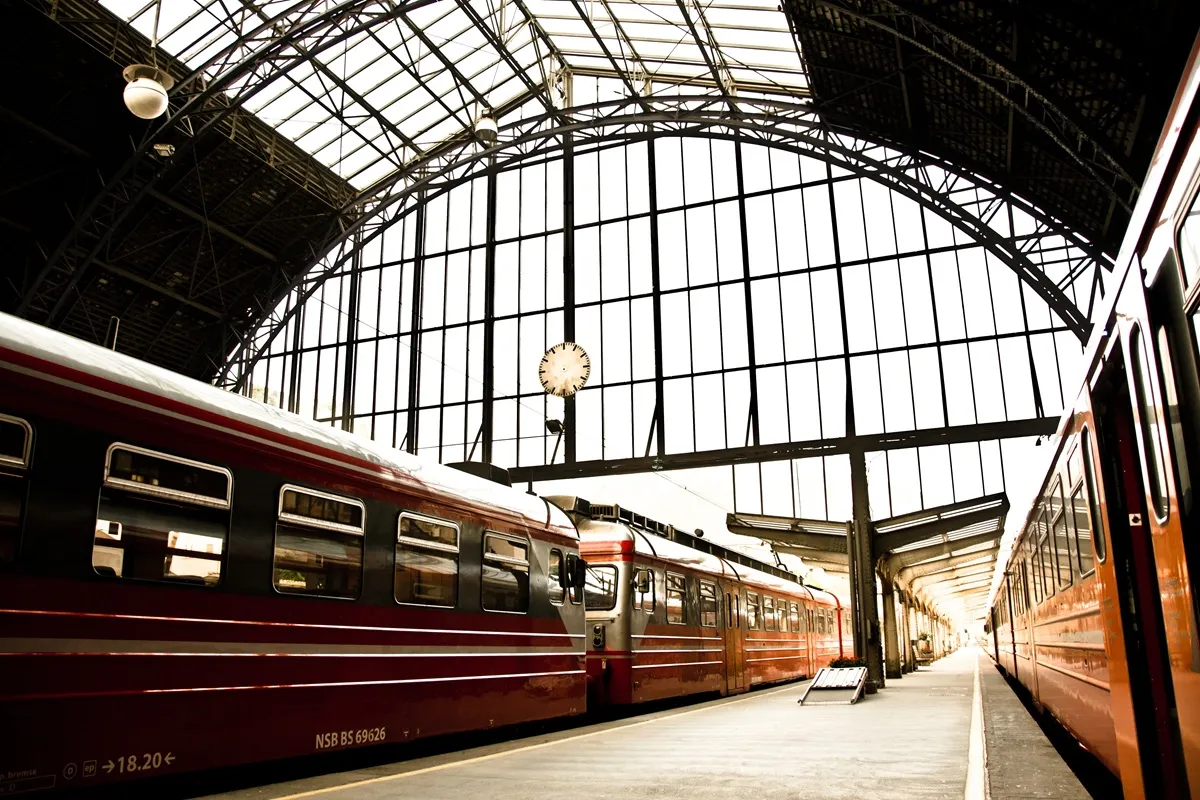 Interno di una stazione ferroviaria storica con treni rossi fermi ai binari sotto una grande volta in vetro e struttura metallica.