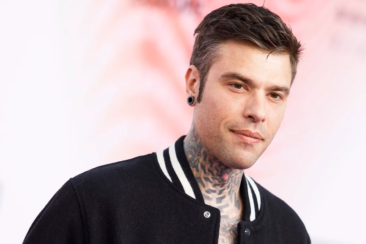 Primo piano di un uomo con capelli corti scuri, tatuaggi geometrici sul collo, orecchino a cerchio nero e giacca sportiva, su sfondo sfumato rosa.